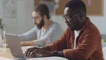 Afro-American businessman using laptop at office desk with glass spit protection wall during coronavirus pandemic while his Arab colleague working in background - Powered by Adobe