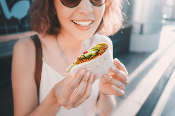Happy woman eats a fast food flatbread with filling sitting on the street and resting after a...