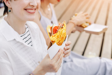Two happy women friends relax in park and eat street food to take away and drink soft drinks. Casual socializing and urban fast food holiday