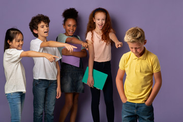 No to bullying. Diverse schoolchildren laughing at sad blonde boy over lilac background