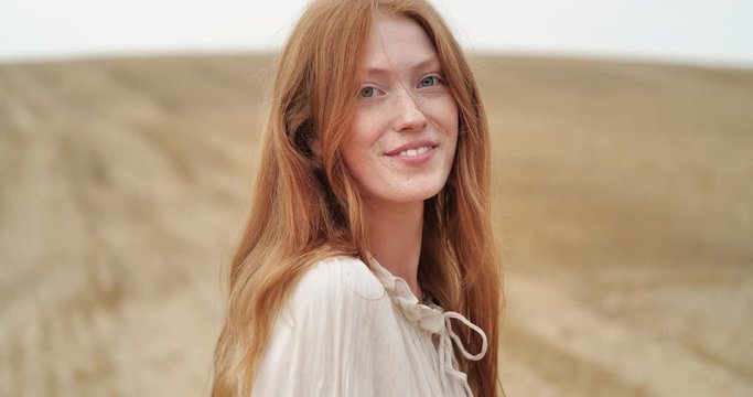 Cute Ginger Woman with long Red Hair walking on the Field, looking at Camera. Happy Ginger Girl is spinning ti the Camera with happy Smile, Summer Face Female. Emotions. People. Portrait.