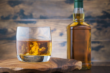 Golden whiskey witn ice cubes on wooden table with bourbon or scotch bottle