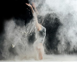 beautiful caucasian woman in a black bodysuit with a sports figure is dancing in a white cloud of flour