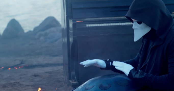 a man in a venetian mask on the seashore plays the glucophone against the background of a burning old piano