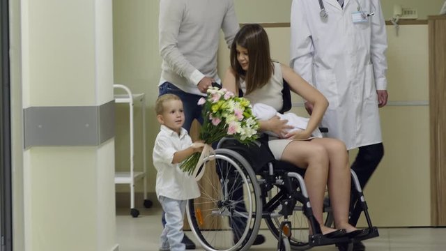 Happy Woman Holding Newborn Baby, Riding Wheelchair With Help Of Husband And Talking With Little Son Holding Bouquet Of Flowers While Leaving Maternity Hospital After Recovery