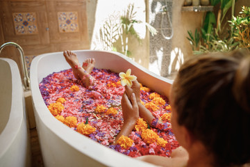 Aromatherapy and spa. Woman lie in bath tube full of flower petals, hold in hands frangipani flower. Body care concept