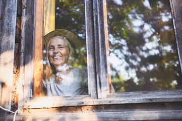 Happy smiling emotional elderly woman having fun posing by open window in rustic old wooden village house in straw hat. Retired old age people concept. Quarantine in the country house.