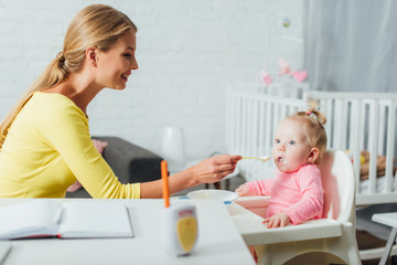 Fototapeta premium Selective focus of mother feeding baby girl on highchair near notebook on table