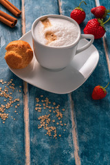 Cup of cappuccino, eclair, strawberries and cinnamon sticks on a background of blue wooden boards.