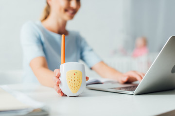 Selective focus of woman holding baby monitor while using laptop at table