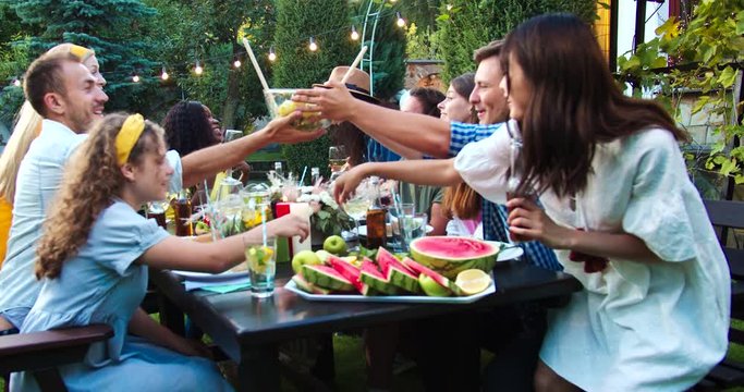 Happy young multi-ethnic people together at garden party. African American and Caucasian girls laughing. Joyful guys and females passing dish while chatting. Pretty Asian girl smiling. Summer concept
