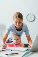 Selective focus of mother working with laptop and writing on notebook while child holding stapler...