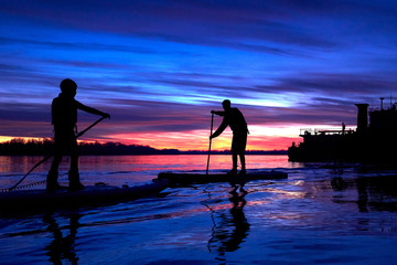 Silhouettes of two little boys paddling on a SUP boards on the autumn Danube river at sunset. Stand...
