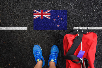 a man with a shoes and travel bag is standing on asphalt next to flag of New Zealand and border 