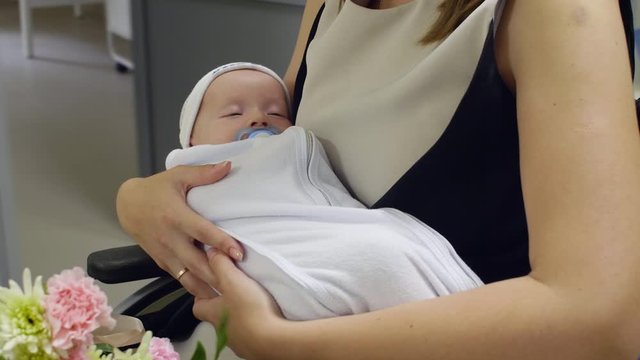 Close up shot of hands of woman holding adorable newborn baby with soother while riding wheelchair through maternity hospital after postpartum recovery