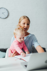 Selective focus of woman using laptop while holding baby daughter at home