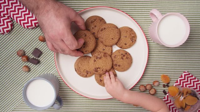 Top view hands take fresh cookies from plate and cup milk. Close up father's and child hands take fresh home cookies. Cute family snack