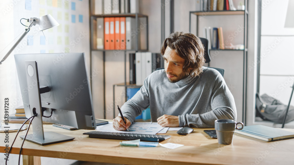 Poster handsome long haired entrepreneur sitting at his desk in the office works on desktop computer, worki