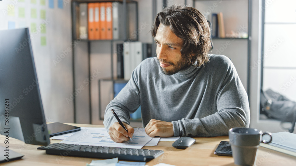 Poster handsome long haired entrepreneur sitting at his desk in the office works on desktop computer, worki