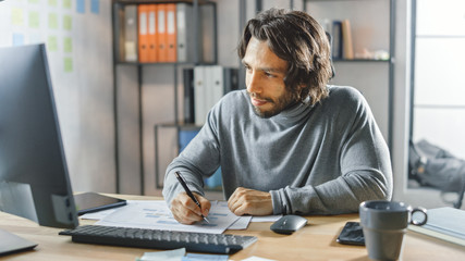 Handsome Long Haired Entrepreneur Sitting at His Desk in the Office Works on Desktop Computer, Working with Documents, Correcting Charts, Drawing Graphs