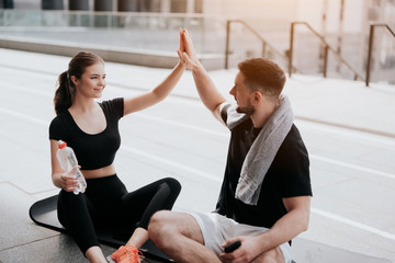 happy handsome brunette man and beautiful woman in sportswear sitting on yoga mat, give each other high five after successful street workout, healthy fit lifestyle