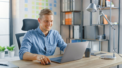 Creative Entrepreneur Sitting at His Desk Works on a Laptop in the Stylish Office, Uses Notebook for Social Media Apps, Emailing Business Associates, Reading News, Browsing through Internet