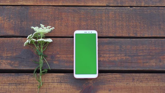 Closeup top view flatlay 4k video footage of modern mobile smartphone with empty blank green screen lying on wooden brown desk background. Woman scrolls green pages. Banner with copy space.