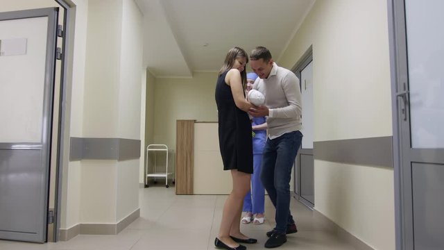 Delightful husband meeting his wife in maternity hospital and taking newborn baby while cheerful nurse giving bouquet of flowers to woman. Loving parents walking together through hallway