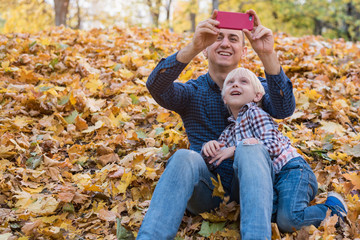 Father and son sitting on autumn leaves and make pictures on phone.