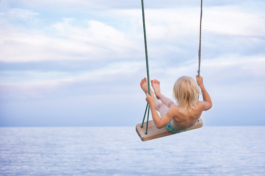 Child Rides A Rope Swing On Sea Background. Vacation At Sea With Children.