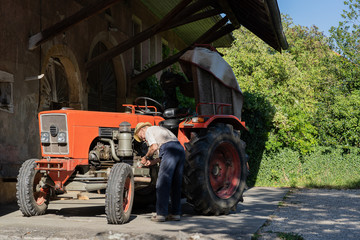 Old farmer working on red tractor under roof of farmer house in sunshine.