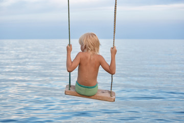Fair-haired boy riding a rope swing over the sea. Summer holidays with children