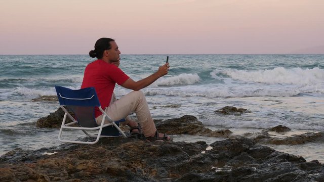 Handsome man stare to phone, sitting in chair on sea beach, evening time. Waves roll and splash at rough stones, neat natural light after sunset. Guy fixed on mobile device