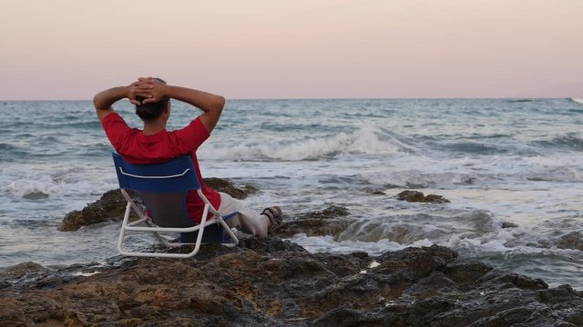 Man stare to sea water, sitting in chair at stony beach, evening time. Slow motion shot, waves slowly roll and break on shallow stones. Peaceful and meditative picture of nature