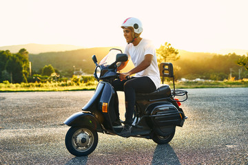 Panoramic of boy on motorcycle with sunset in the background. Dark-haired boy with white helmet with flag of Italy with white t-shirt and black motorcycle with mountains in the background.