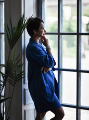 Thoughtful short haired brunette woman in modern blue suit and white stands at floor-to-ceiling window and looks outside