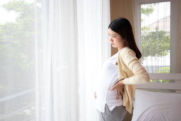 Young beautiful pregnant asian woman standing and touching belly and back near the window in bedroom. Pregnant women with low back pain, stress, anxiety concept.