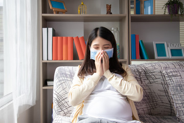 Pregnant asian female wearing mask mouth, She sitting on sofa and feels sick and coughing. Pregnant woman belly closeup protective medical mask from coronavirus, air pollution.