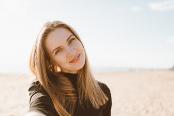 Pretty female with long hair takes selfie on smartphone on beach in summer or autumn. Beautiful...