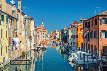 Fototapeta na wymiar Chioggia cityscape with narrow water canal Vena with moored multicolored boats between old colorful buildings and brick bridge, blue sky background in summer day, Veneto Region, Northern Italy