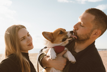 Young happy couple with dog taking selfie photo on beach. Beautiful family and Corgi puppy lick man cheek. Family with pet outdoor, walking together, husband and wife relax in summer by sea