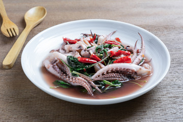 close up of stir fried squid tentacle with holy basil in a ceramic dish on wooden table. hot and spicy thai style food menu.