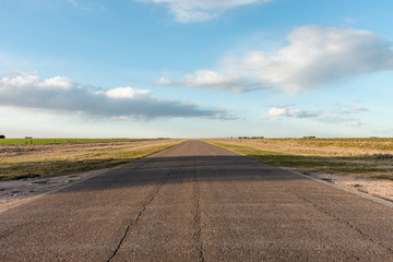 Empty asphalt road going through agricultural fields in countryside zone. Light blue sky with soft clouds at sunset golden hour.