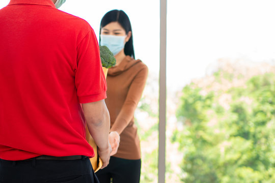 Asian Woman Wearing A Face Mask And Pickup Shopping Bag Of Fresh Food, Vegetables, And Fruits From A Delivery Man From A Supermarket. Concept Of Express Grocery Service And New  Lifestyle