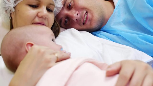 Close up tilt up shot of joyous mother and dad in medical hats lying together on couch in hospital ward, smiling and caressing newborn baby