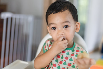 An Adorable toddler Asian baby boy (1-year-old) sitting on high feeding chair eating homemade food...