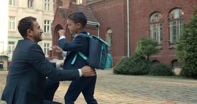 Little cheerful kid embracing his loving father and waving while running at lessons. Young man in suit watching his son going to school. Concept of family and education.