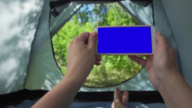 Closeup point of view 4k video shoot of two white female hands holding modern mobile phone while laying inside of camping tent in sunny summer forest. Blank blue screen of smartphone for your design.