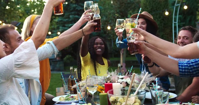 Portrait of joyful mixed-races young friends raising glasses and clinking. Caucasian males, Asian and African American girl chatting gathered together at party at table in garden. Summer fun concept