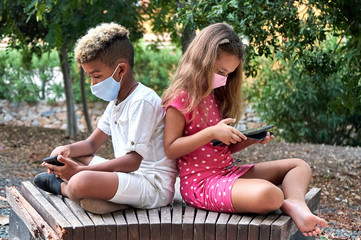 Two school age 9s 10s multi racial kids wear face mask sit together on bench back to back use cellphone and tablet devices ignore each other. Phubbing, new generation addicted with modern tech concept